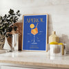 Aperol cocktail poster in bold blue and yellow modern abstract illustration - styled on a counter with eucalyptus and a bowl of lemons