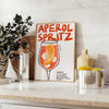 Poster raffigurante un Aperol Spritz stilizzato in arancione e nero - su un piano con eucalipto e una ciotola di limoni