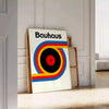 Poster showing Bauhaus geometric artwork with bold shapes and modern composition - leaning on a herringbone floor against a white panelled wall
