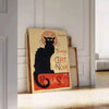 A vintage poster featuring a black cat with a yellow background, text reading 'Chat Noir'. - leaning on a herringbone floor against a white panelled wall