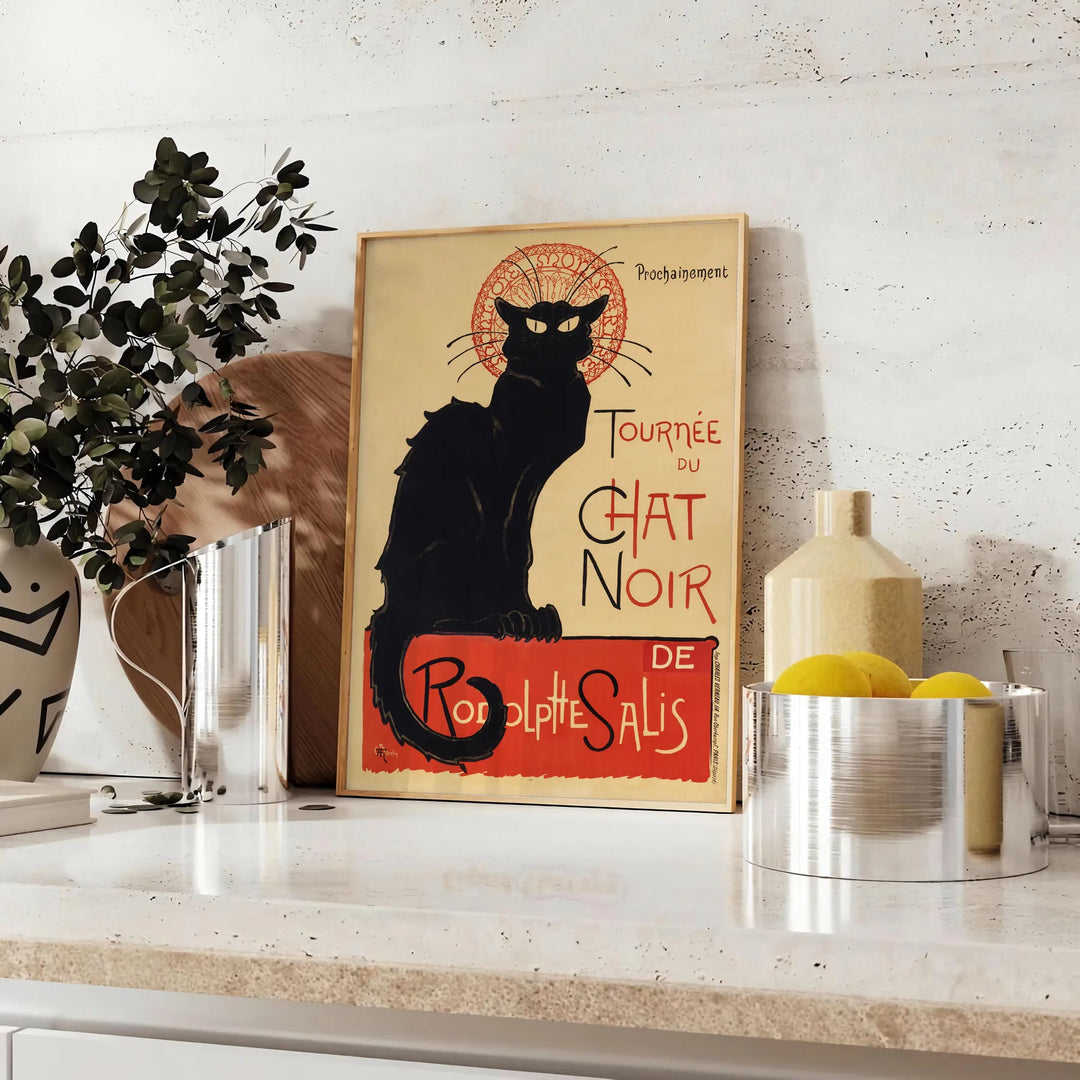 A vintage poster featuring a black cat with a yellow background, text reading 'Chat Noir'. - styled on a counter with eucalyptus and a bowl of lemons