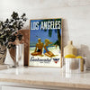 Vintage travel poster featuring Los Angeles with green palm trees and white lettering - styled on a counter with eucalyptus and a bowl of lemons