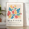 Poster showing colorful abstract paper cutouts in Matisse style artwork - in a bright minimalist room with a plant and an armchair