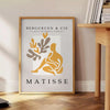 Poster showing Matisse-style papier découpés with bold colorful abstract shapes - beside a round wooden side table with books