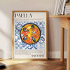 Eine farbenfrohe Darstellung eines Paella-Gerichts mit warmen Orange- und Weißtönen - neben einem runden Beistelltisch aus Holz mit Büchern
