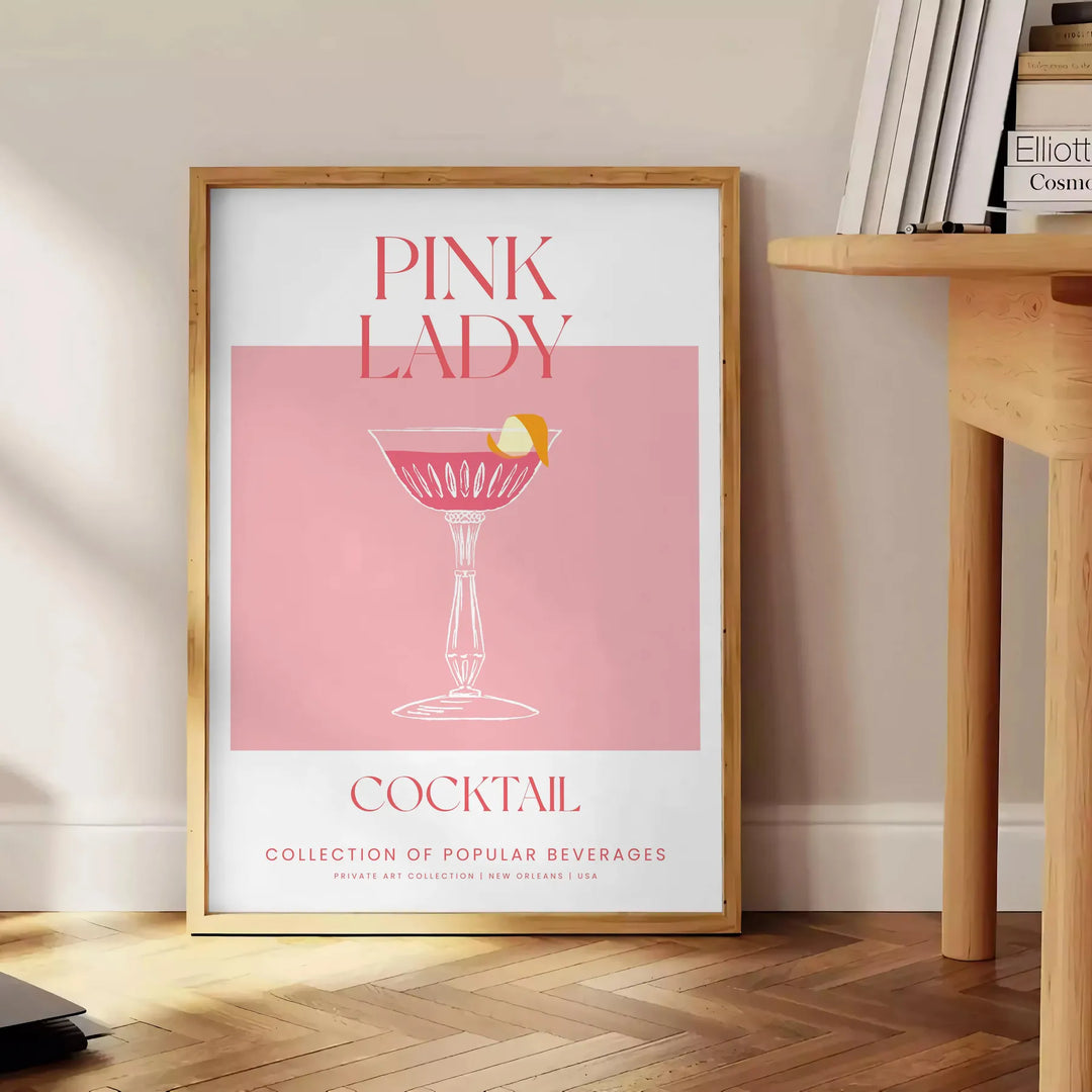 Kolorowa ilustracja koktajlu Pink Lady w odcieniach pomarańczowym i różowym - obok okrągłego drewnianego stolika z książkami