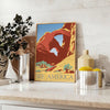 A retro travel poster featuring a yellow and blue landscape with mountains and a road - styled on a counter with eucalyptus and a bowl of lemons