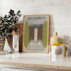 A vintage travel poster featuring the text 'See America' with green and gray landscapes. - styled on a counter with eucalyptus and a bowl of lemons
